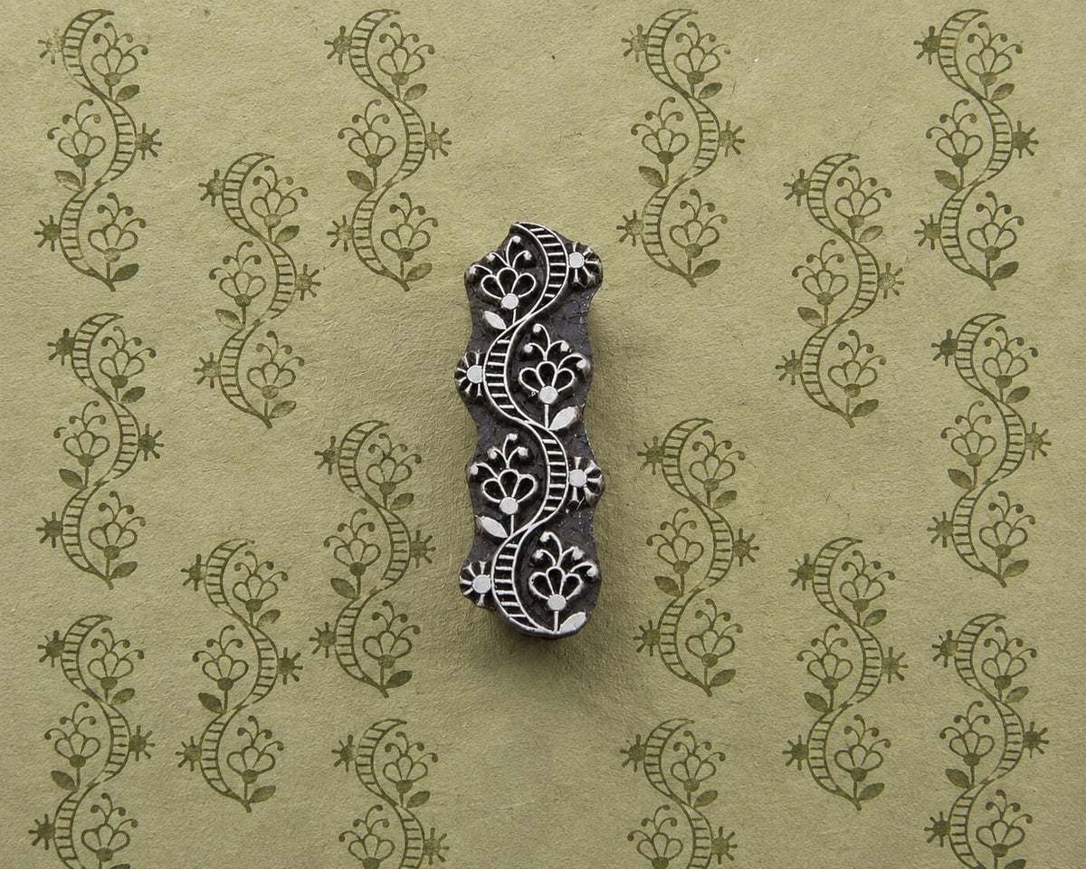 Hand-carved wooden printing block with a flowing floral ornament, shown with repeated green impressions on textured handmade paper.