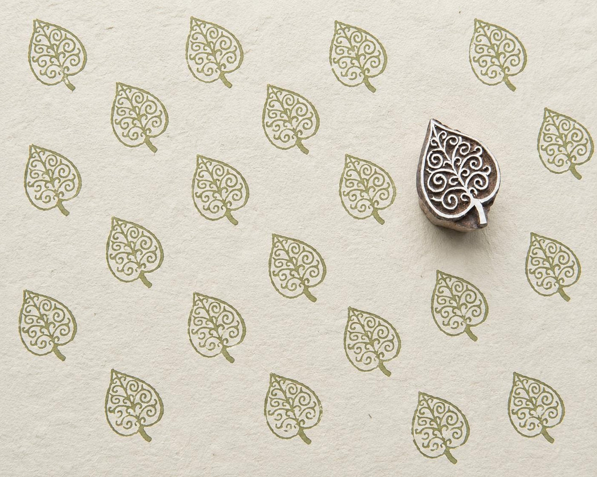 Hand-carved wooden printing block with an intricate scrolling leaf motif, shown with repeated olive-green impressions on handmade paper.
