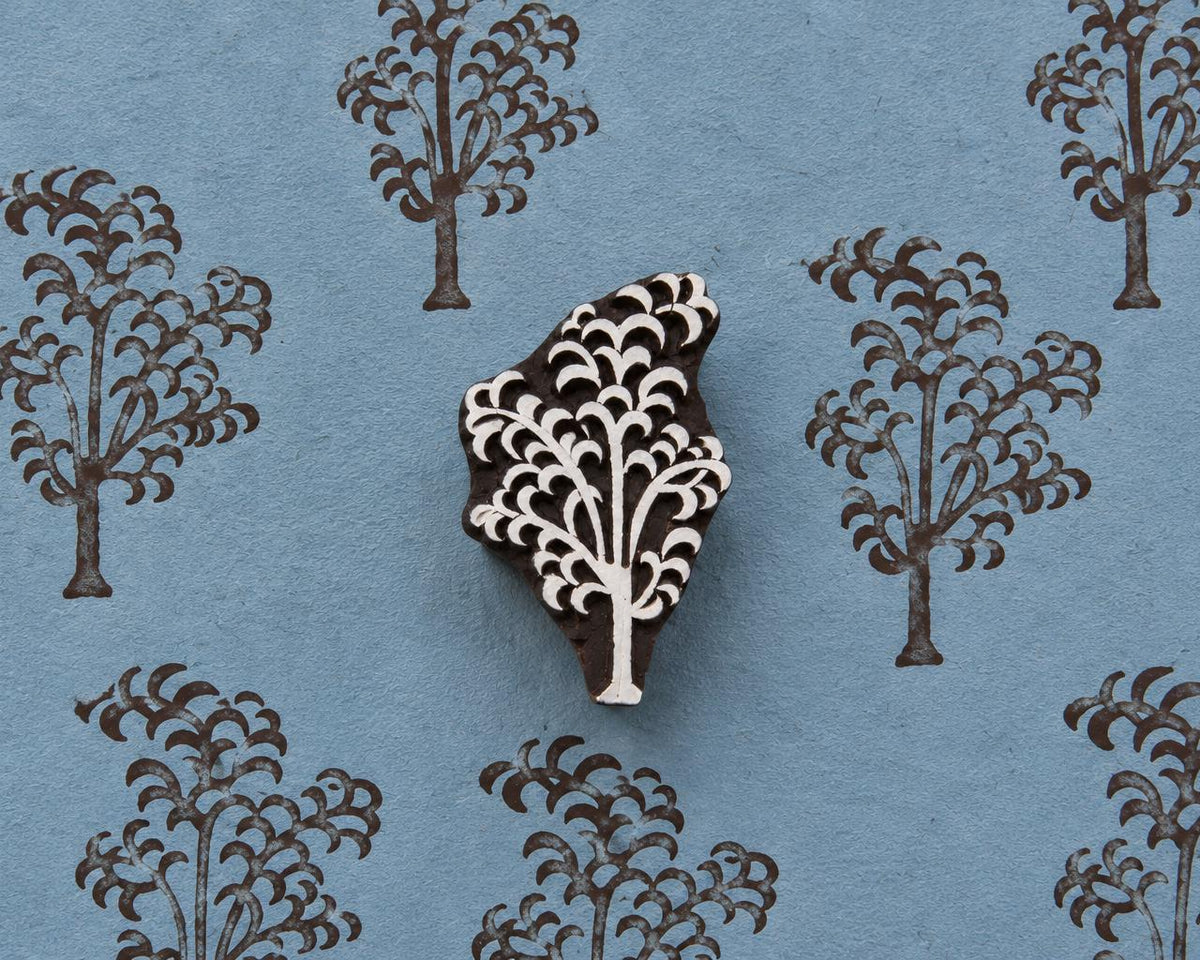 Hand-carved wooden printing block of a tree with a cascading branch and curved foliage motif, shown with repeated brown impressions on blue handmade paper.