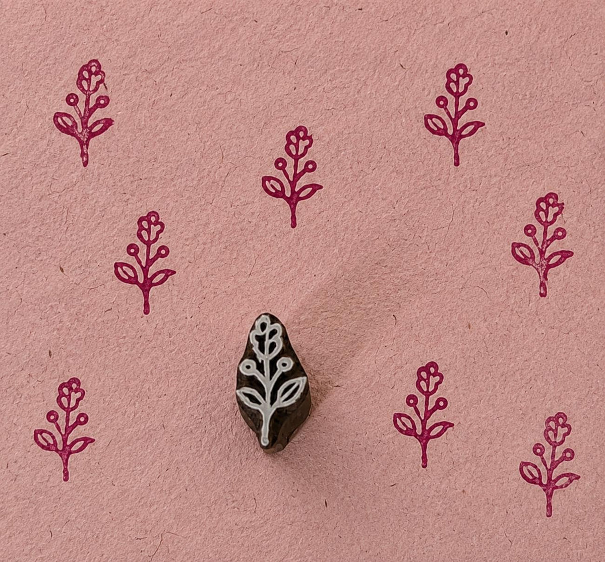 Hand-carved tiny wooden stamp featuring a delicate Berry Blossom Sprig, shown with repeated burgundy impressions on textured handmade pink paper.