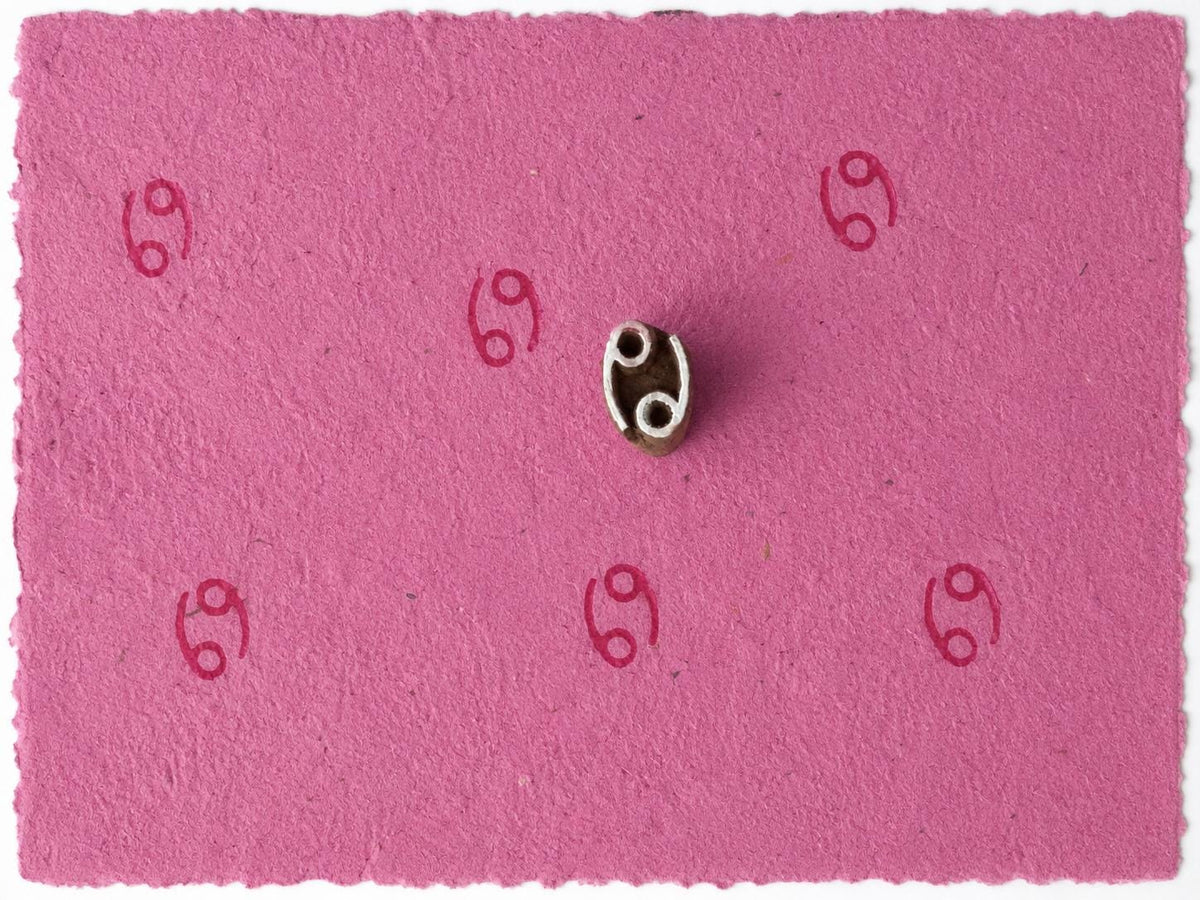 Tiny Cancer zodiac symbol stamped repeatedly on pink handmade paper with wooden printing block