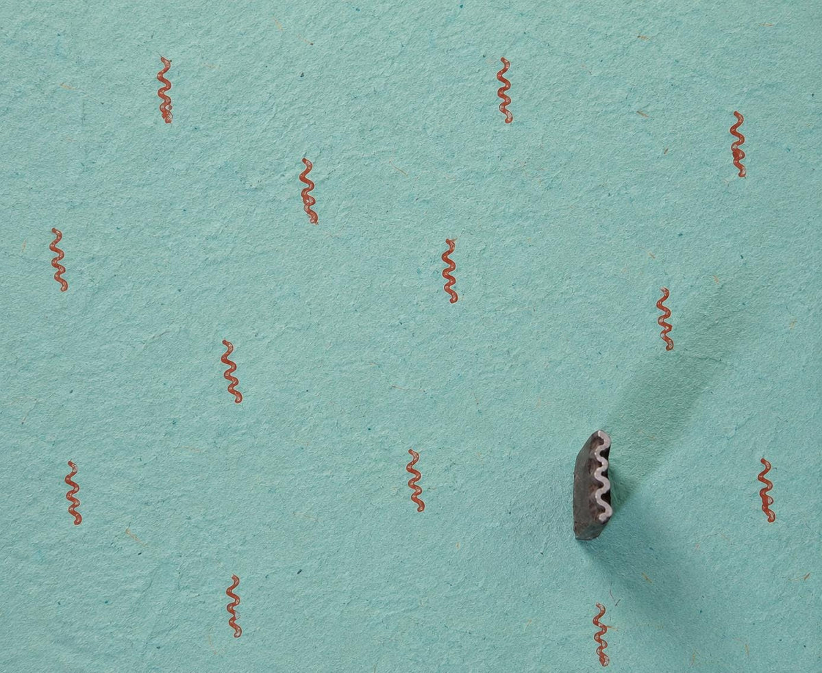Tiny wavy vine motif repeatedly printed in rust ink on textured turquoise handmade paper with the carved wooden block shown alongside.