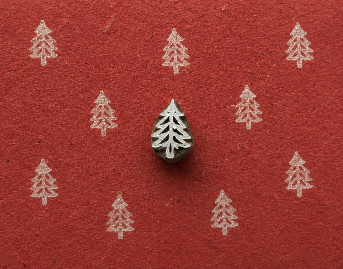 Simple Evergreen wooden stamp shown with repeated white tree impressions on textured red handmade paper.