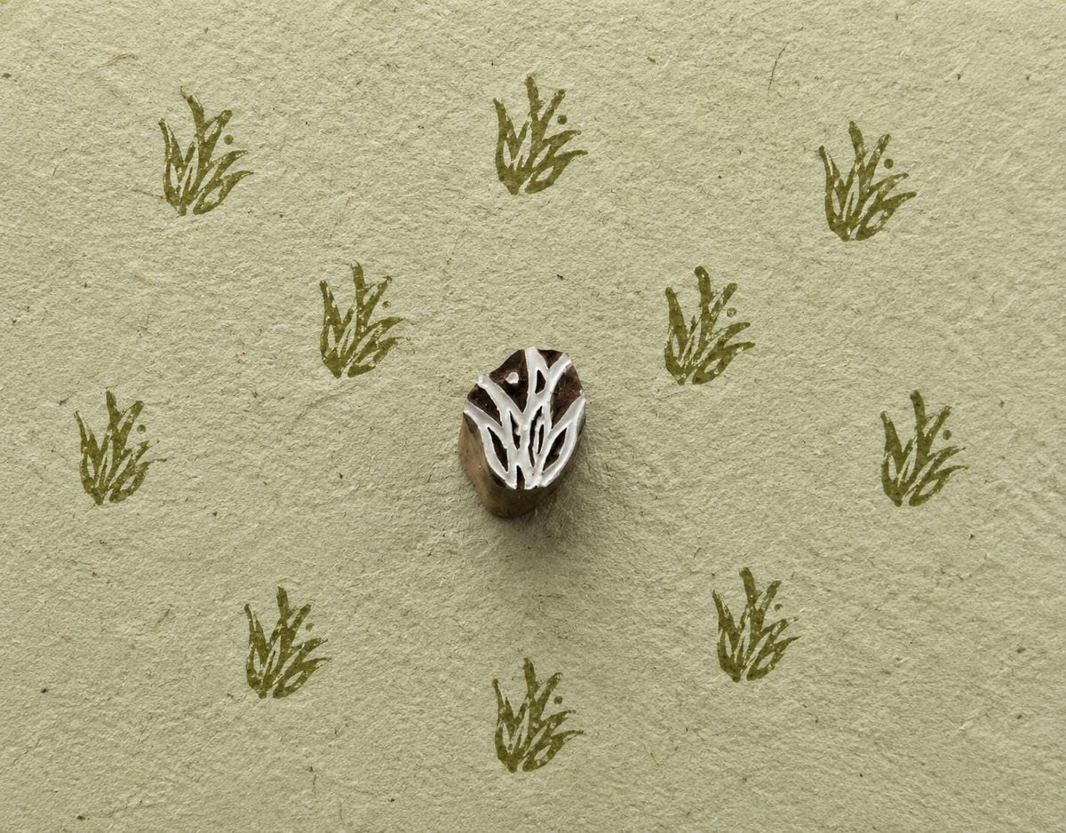 A delicate botanical motif inspired by a small tuft of wild grass, hand carved into wood with fine, organic lines and tiny dot details.