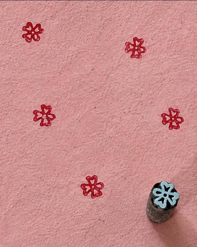 Hand carved Indian wooden printing block with a small petal floral motif, shown creating a repeating red pattern on handmade pink paper.