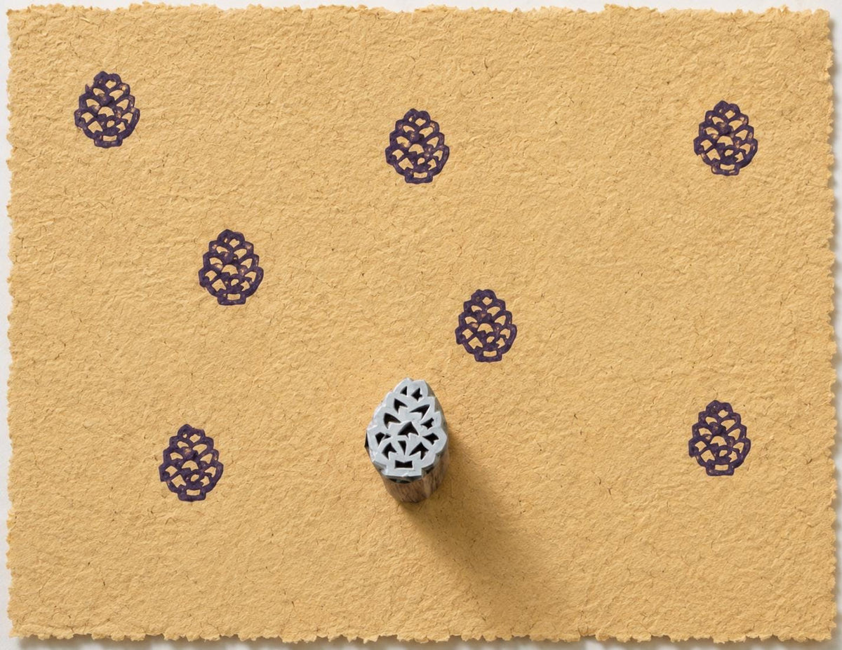 Tiny hand carved Indian wood printing block with pinecone motif, shown with repeated purple impressions on handmade paper