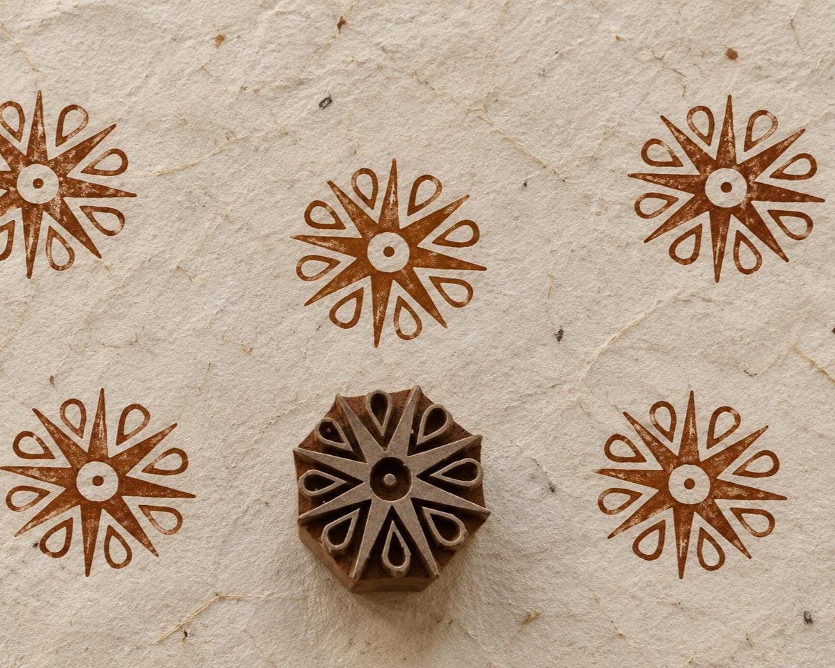 Hand carved wooden printing block with a stylised teardrop snowflake design, surrounded by repeated brown snowflake impressions on textured handmade paper.