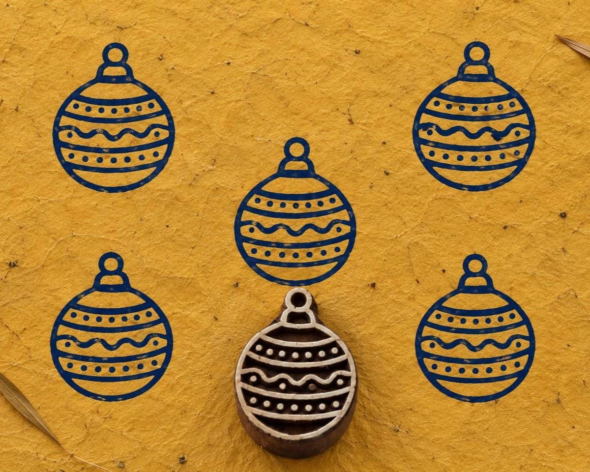 Hand carved festive Christmas ornament wooden printing block surrounded by navy blue ornament prints on textured mustard yellow handmade paper