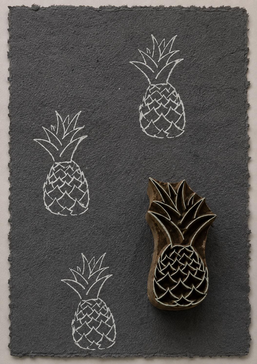 Hand carved tropical pineapple wooden printing block with repeated white impressions on dark handmade paper