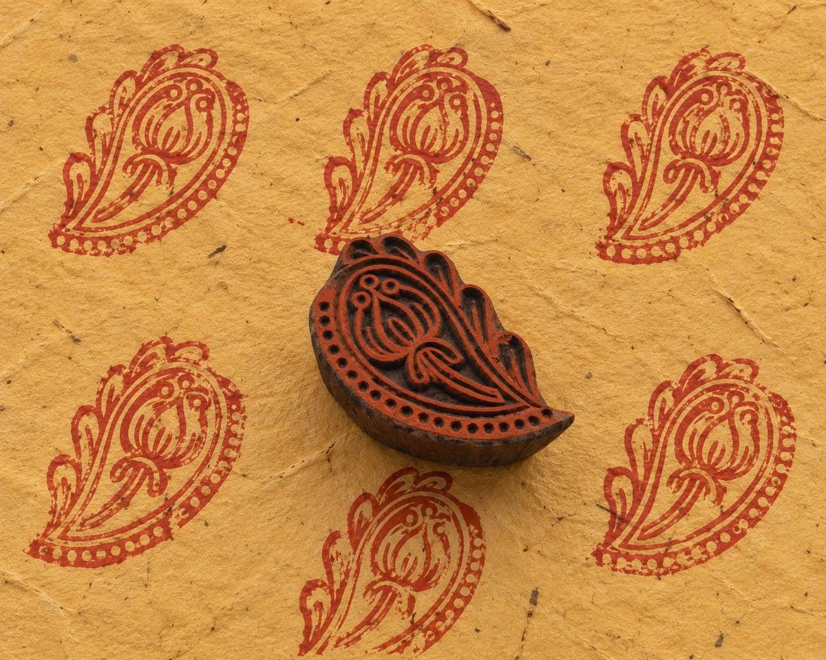 Hand carved Indian wooden printing block with an ornamental floral paisley design, shown beside repeated red prints on handmade paper.