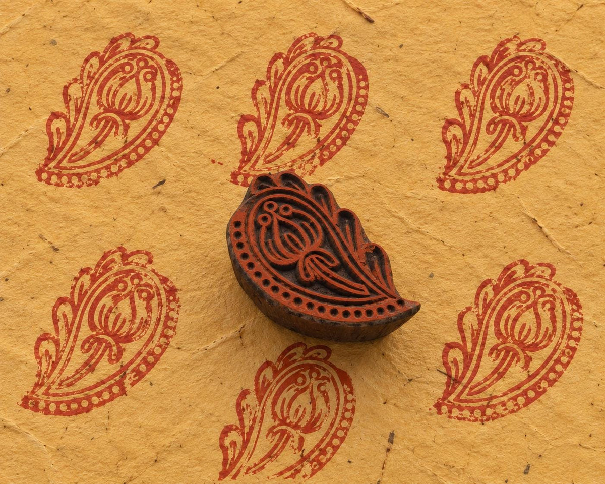 Hand carved Indian wooden printing block with an ornamental floral paisley design, shown beside repeated red prints on handmade paper.