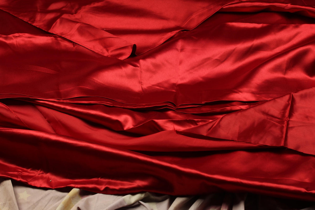 Deep crimson silky faux satin fabric arranged in soft folds showing its fluid drape and glossy texture.