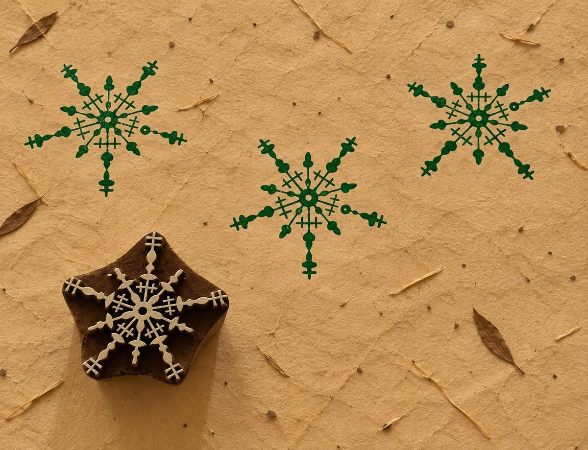 Intricate wooden snowflake printing block with a detailed symmetrical design, shown with repeated green prints on handmade paper.
