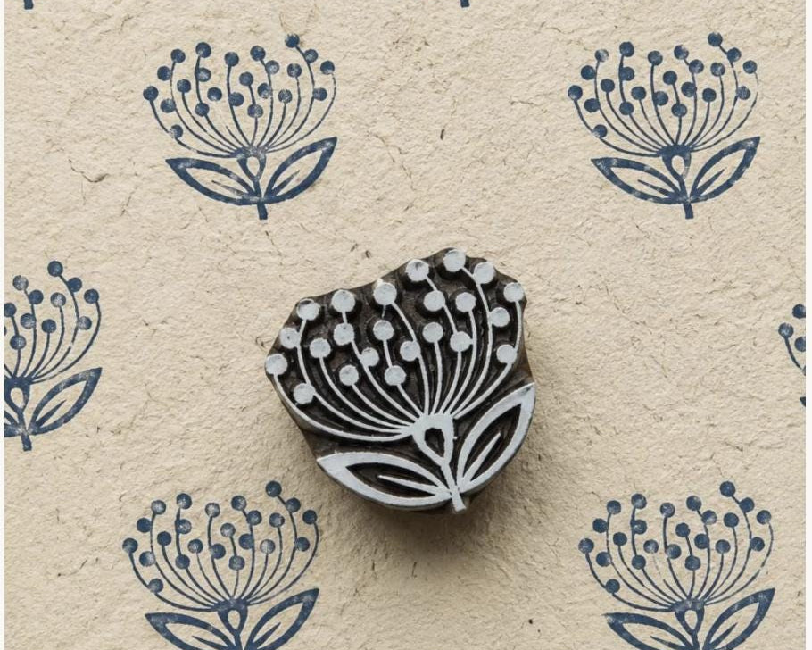 Hand carved wooden dandelion-inspired motif shown with repeated blue impressions on textured handmade paper.