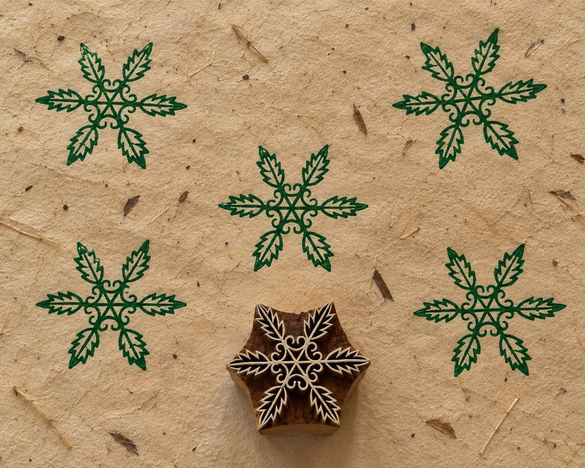 Hand carved wooden printing block featuring a six-leaf botanical snowflake design, with green printed impressions on textured handmade paper.