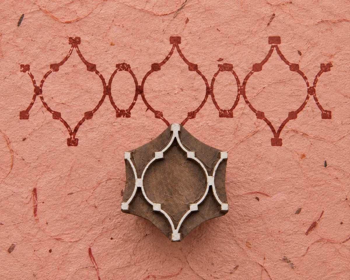 Hand carved wooden geometric tile printing block beside a repeating interlocking border pattern printed in burgundy on textured pink handmade paper.