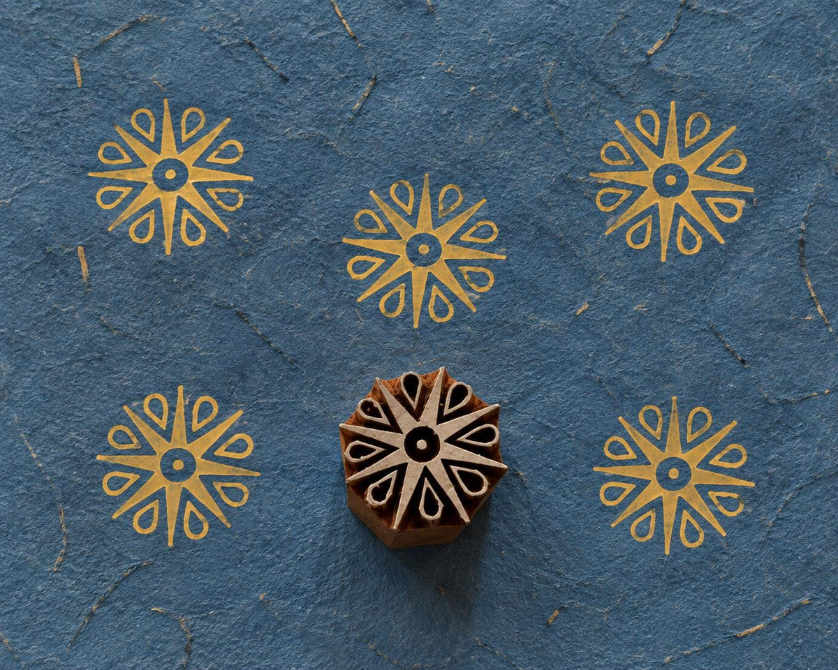 Decorative wooden printing block featuring a simple radial petal motif, shown with repeated golden prints on blue handmade paper.