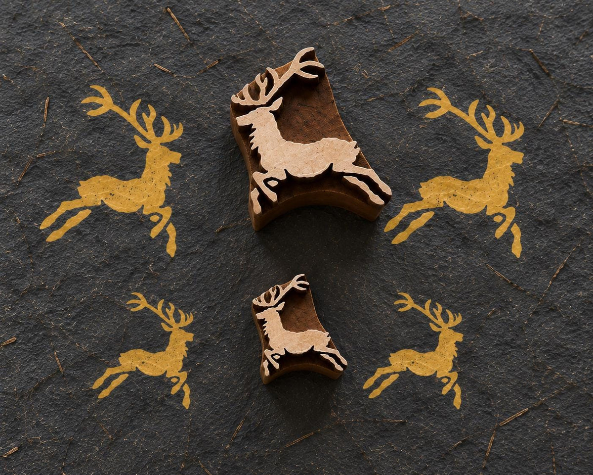 Handmade paper showing repeated brown ink impressions of a whimsical Christmas reindeer set of designs beside its carved wooden printing block.