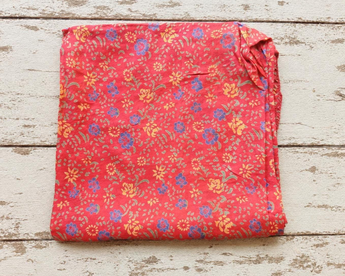 Close view of coral-red fabric with scattered blue flowers, yellow blossoms and green decorative motifs.