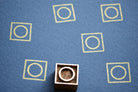 Handcrafted wooden printing block with a circle inside a square, shown on blue paper with repeating yellow stamped patterns