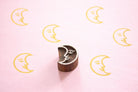 Hand carved Indian wooden printing block with a crescent moon face design shown on a pink surface with repeated yellow moon prints.