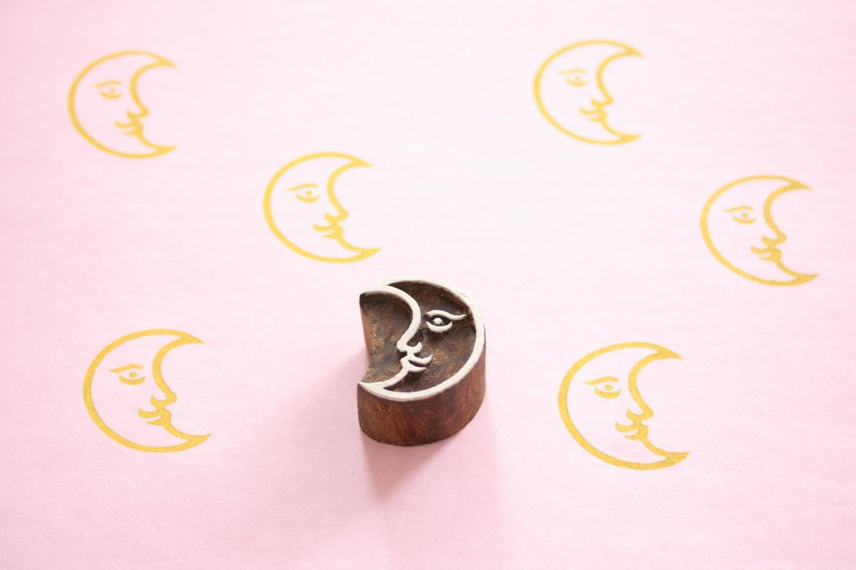 Hand carved Indian wooden printing block with a crescent moon face design shown on a pink surface with repeated yellow moon prints.