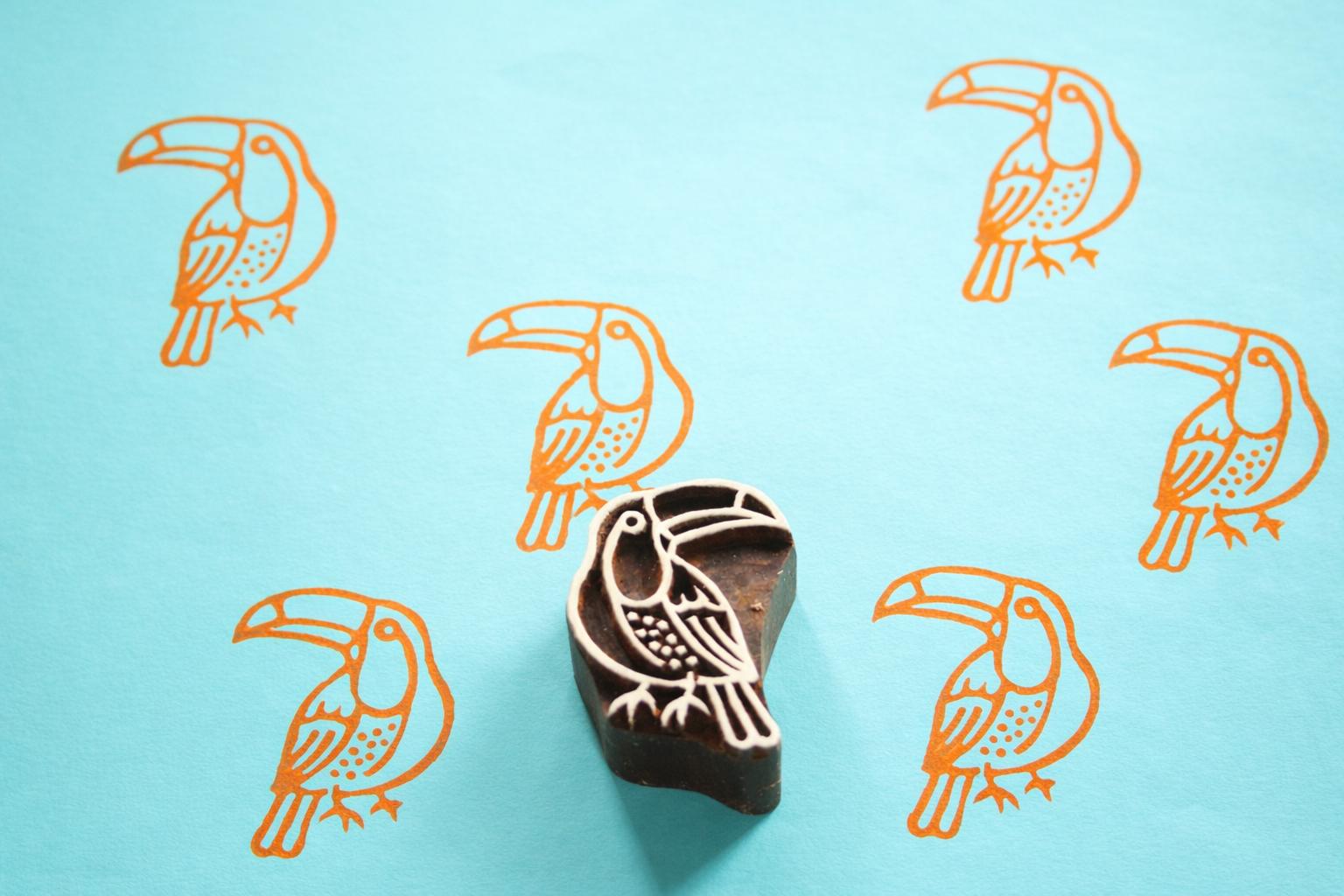 Hand carved Indian wooden printing block with a toucan bird design shown on a blue surface with repeated orange toucan prints.
