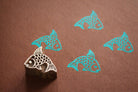 Hand carved Indian wooden printing block with a fish design shown on brown paper with repeated turquoise fish prints.