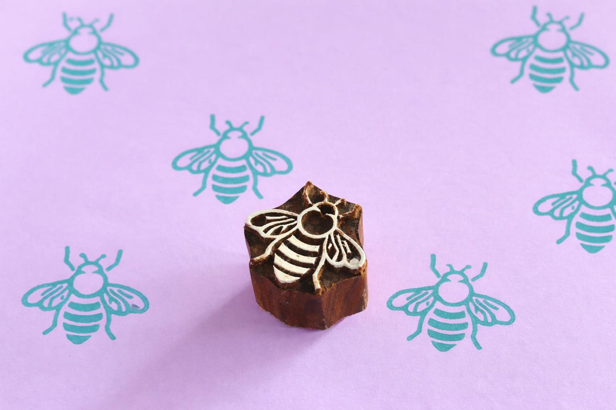 Hand carved bee wooden printing block shown with repeated bee prints on pastel purple paper