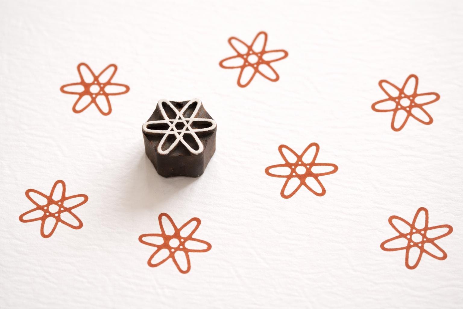 Handcrafted wooden printing block with an atom-style star design, shown on white paper with repeating rust-colored stamped patterns