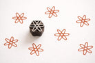Handcrafted wooden printing block with an atom-style star design, shown on white paper with repeating rust-colored stamped patterns