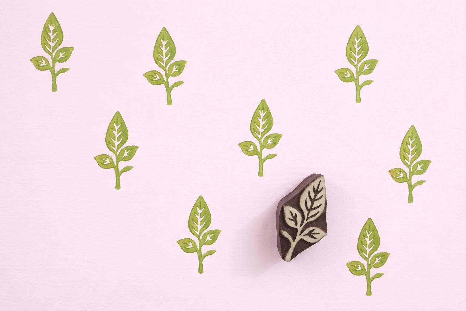 Hand carved oval wooden leaf stem block printing stamp creating light green repeating botanical pattern on soft pink background.