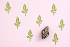 Hand carved oval wooden leaf stem block printing stamp creating light green repeating botanical pattern on soft pink background.