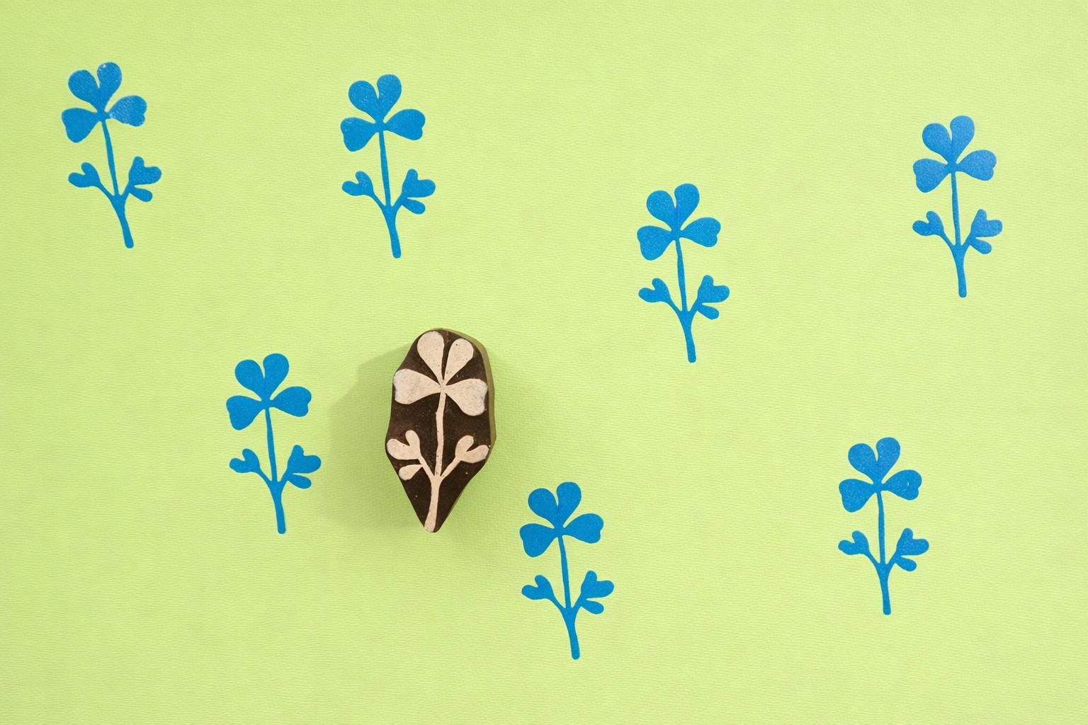 Three-petal floral wooden block printing stamp creating repeating blue botanical pattern on green background.