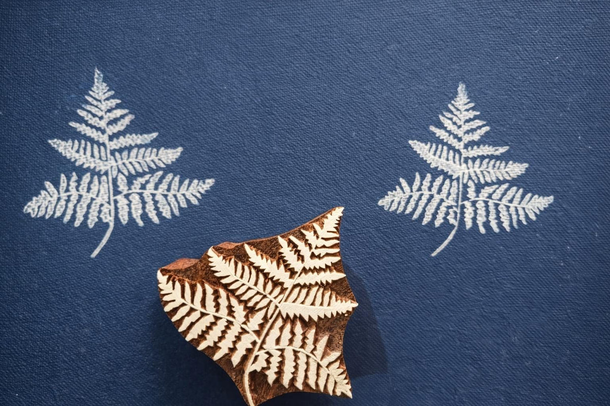 Hand carved wooden fern frond block printing stamp creating detailed white fern pattern on navy blue fabric background.