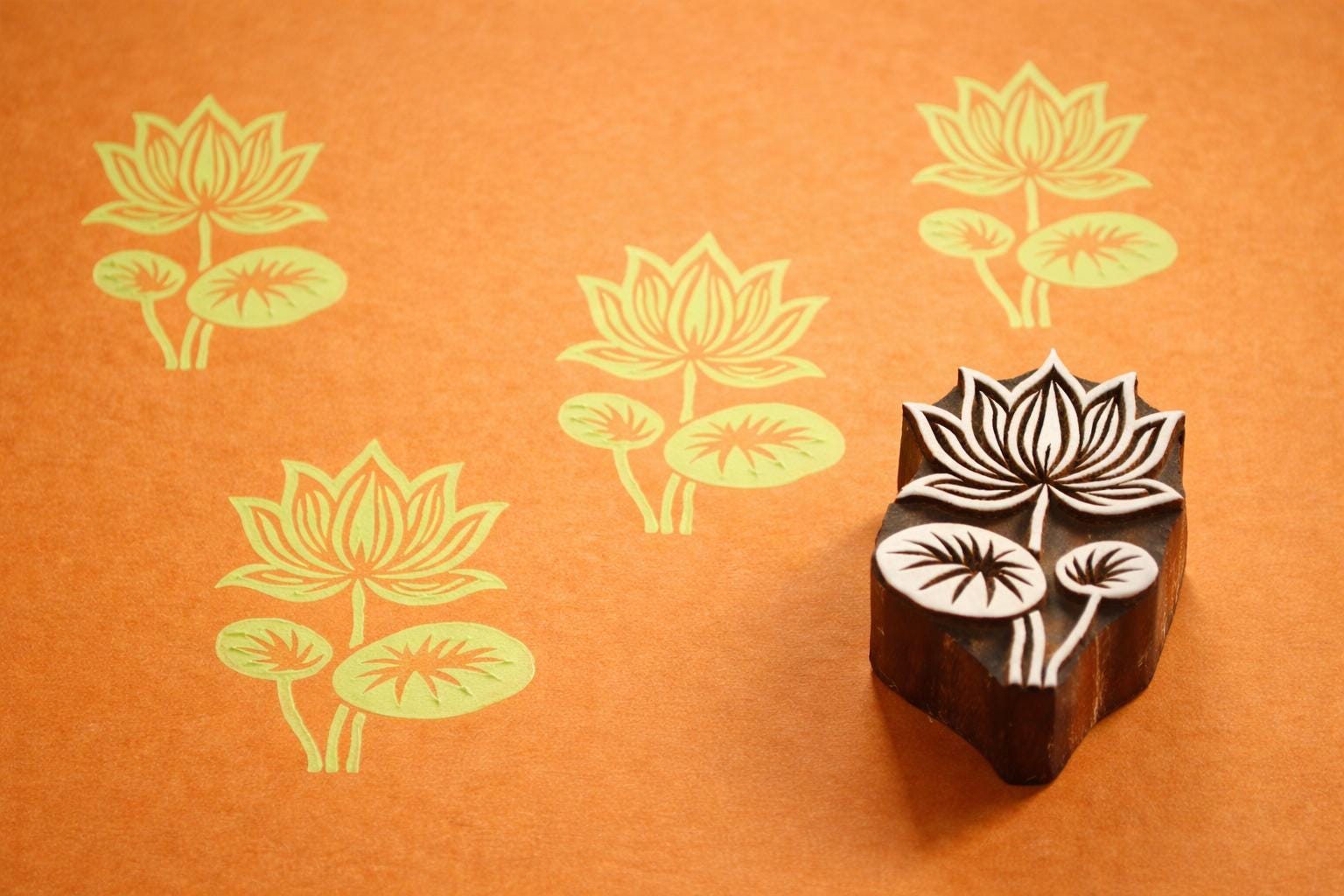 Hand carved Indian wooden printing block with a detailed lotus flower design shown on orange paper with repeated lotus prints.