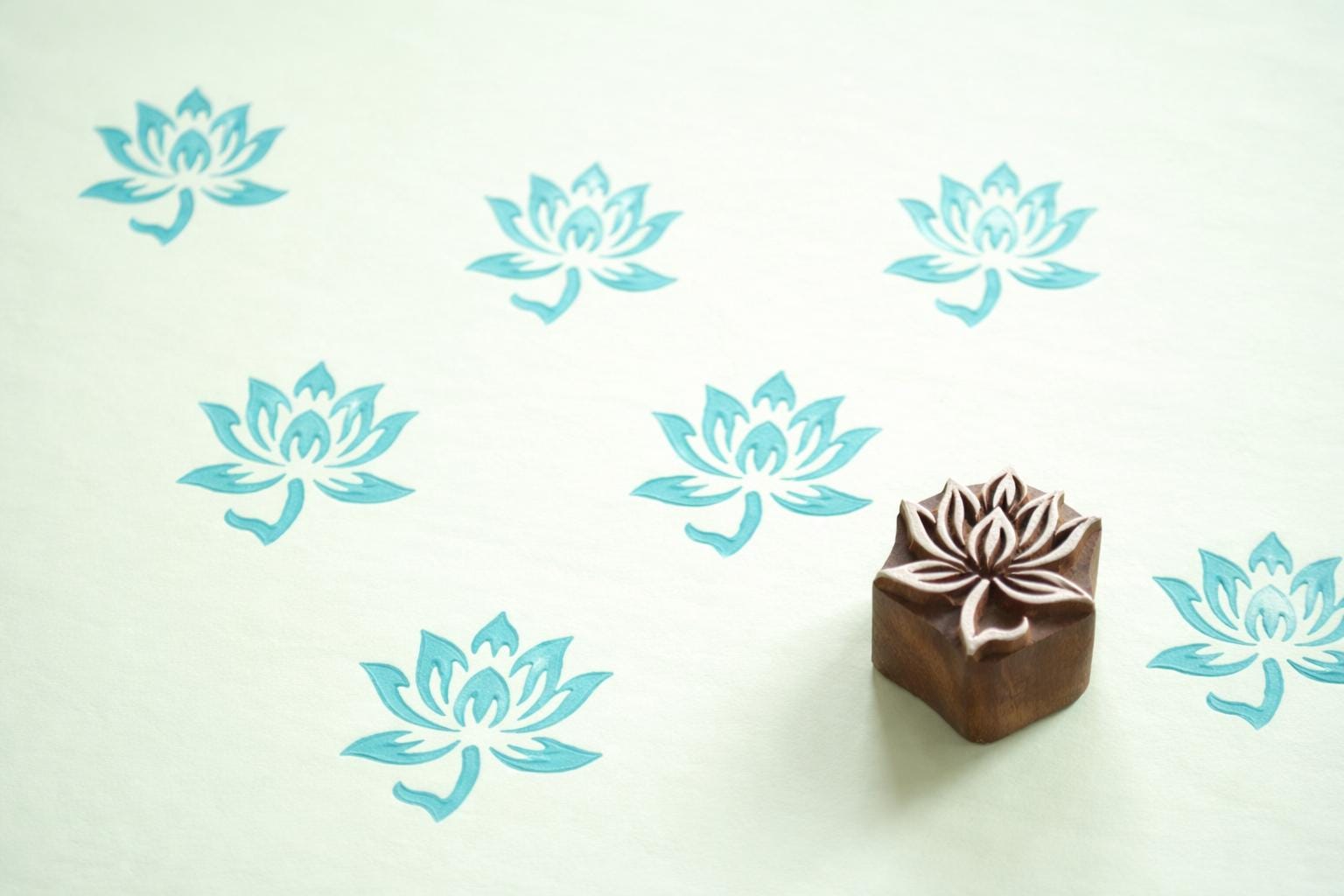 Hand carved Indian wooden printing block with a simple lotus flower design shown on light green paper with repeated blue lotus prints.
