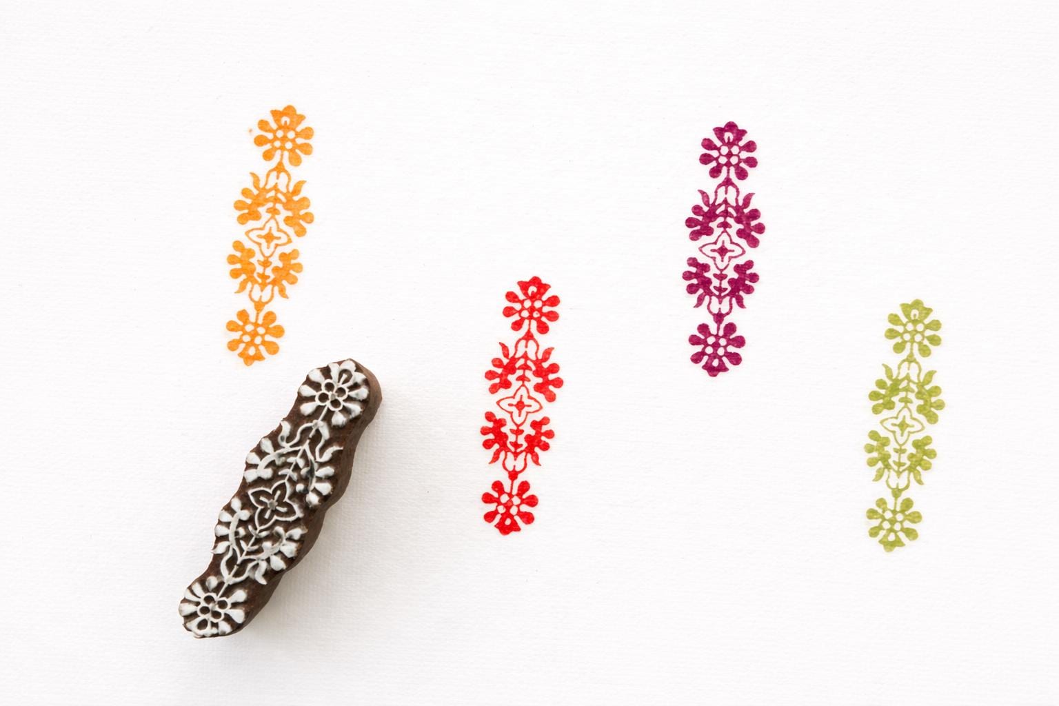 Hand carved wooden floral border block printing stamp creating vertical repeating pattern in multiple colors on white background.