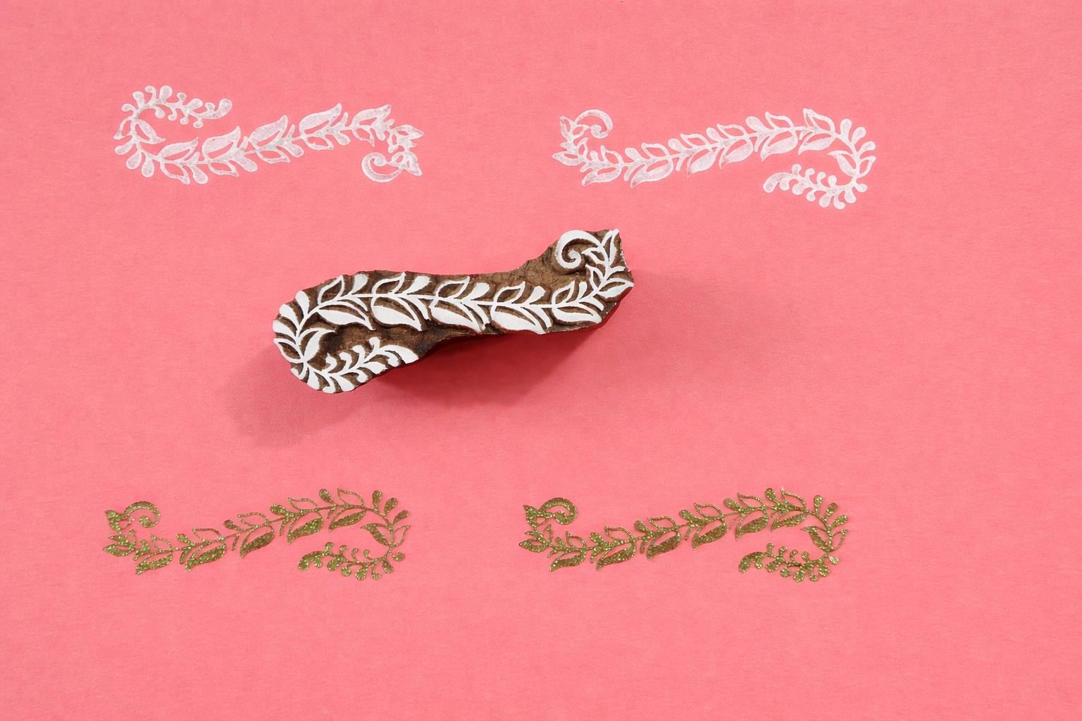 Hand carved wooden curved vine border block printing stamp creating delicate repeating botanical pattern on pink background.