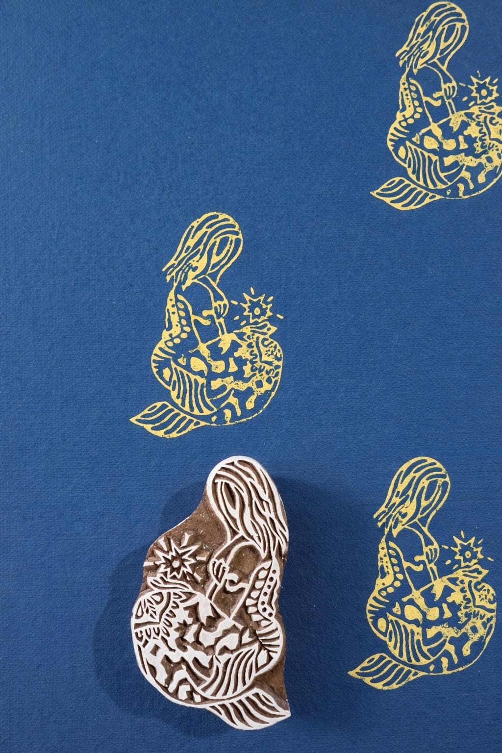 Hand carved wooden mermaid block printing stamp creating yellow ink impressions on blue paper.