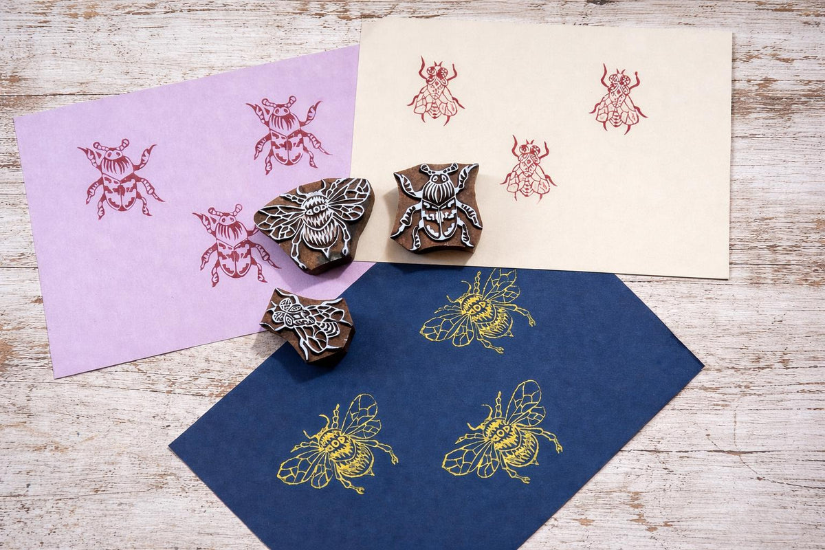 Three hand carved wooden insect block printing stamps shown with bee and beetle prints on colored paper backgrounds.