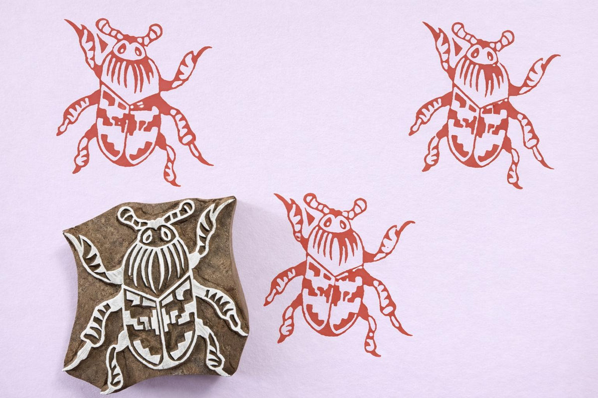 Hand carved wooden beetle block printing stamp creating coral insect prints on pastel purple paper.