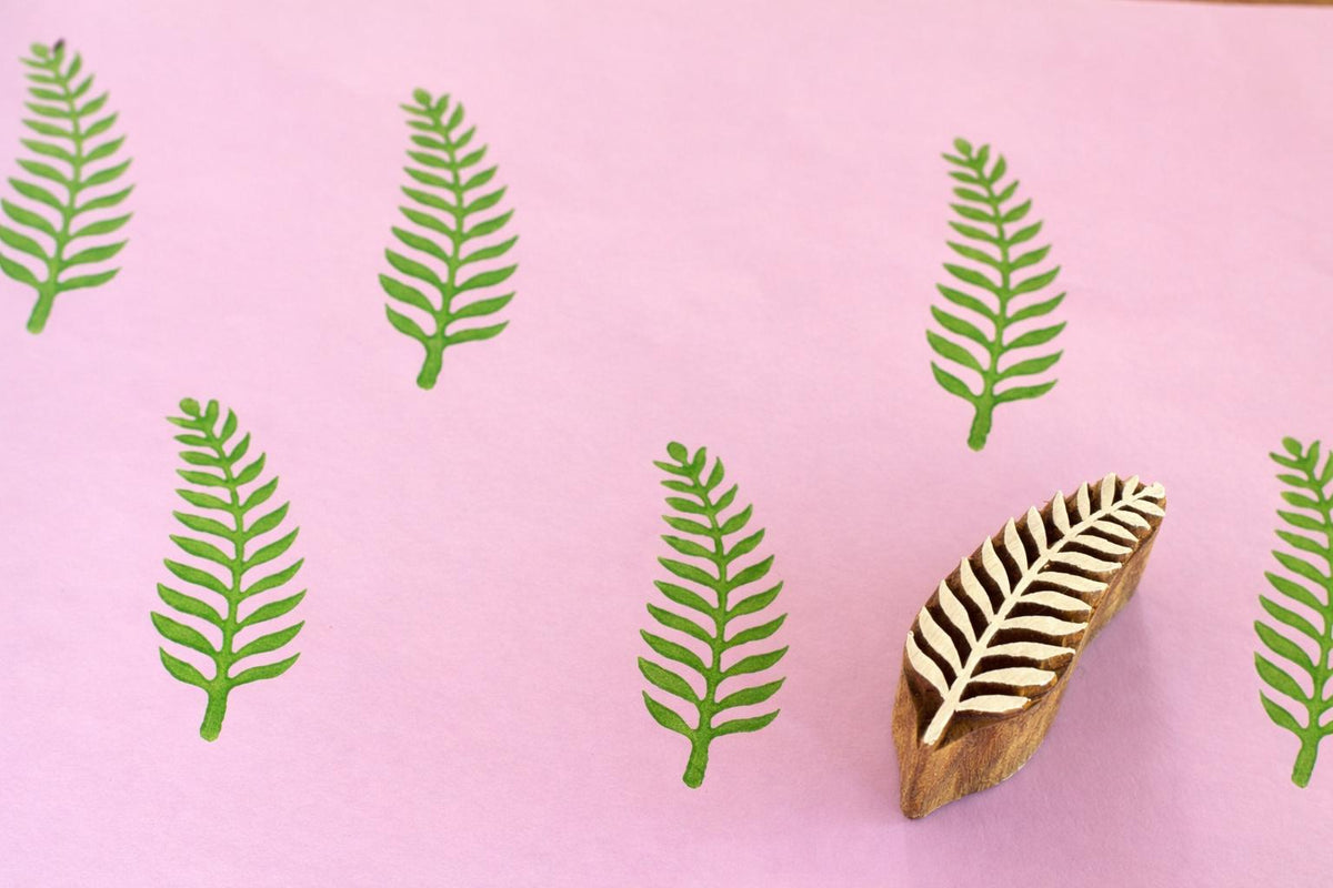 Hand carved fern leaf wooden printing block with green repeated print on pink background.