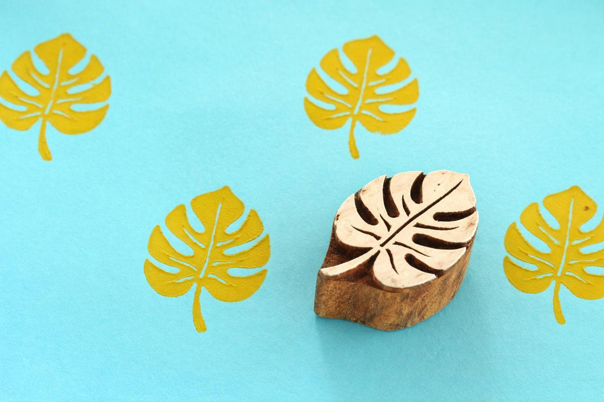 Hand carved monstera leaf wooden stamp with yellow printed pattern on blue background.