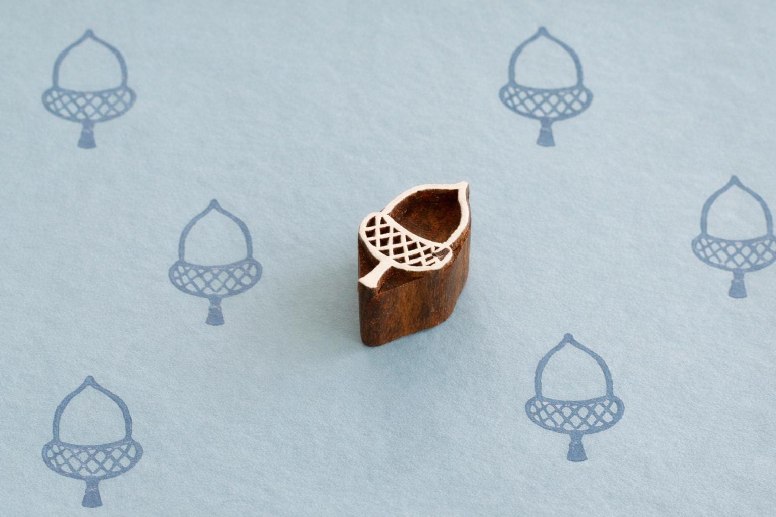 Hand carved wooden acorn printing block stamp shown with blue acorn prints on faded blue background.