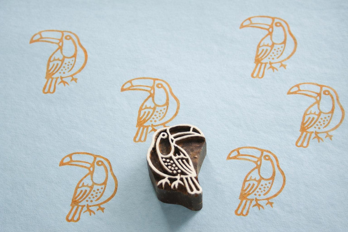 Hand carved Indian wooden printing block with a toucan bird design shown on a faded blue surface with repeated orange toucan prints.