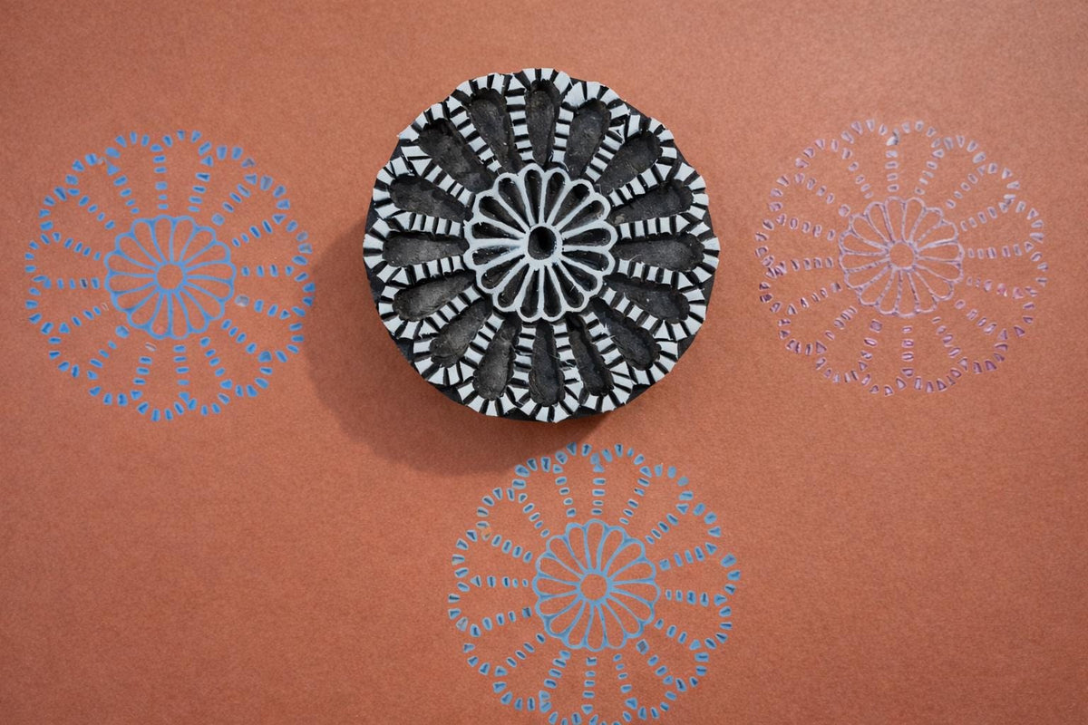 Hand carved round wooden mandala block printing stamp creating blue and purple radial floral pattern on orange background.