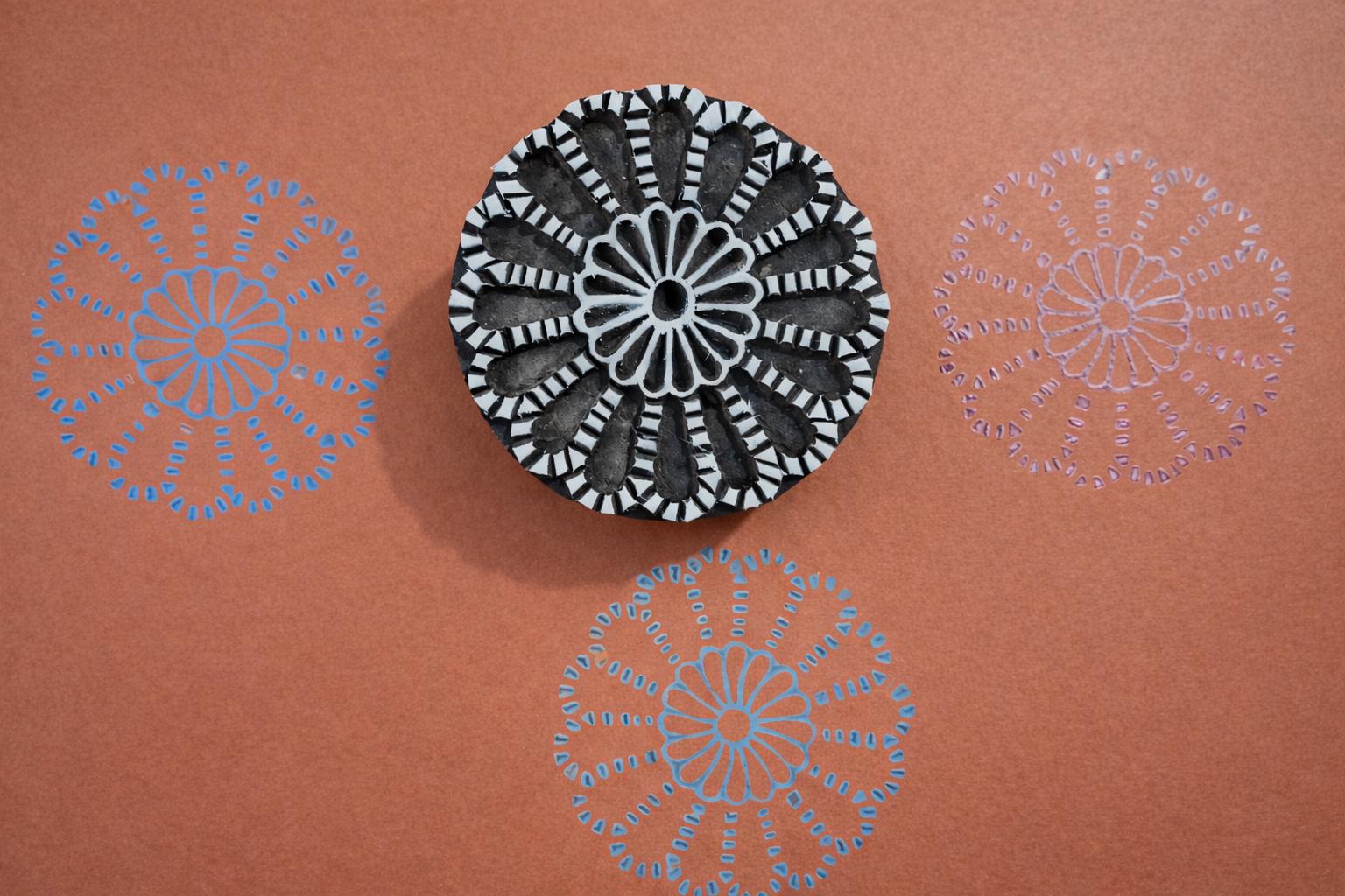 Hand carved round wooden mandala block printing stamp creating blue and purple radial floral pattern on orange background.