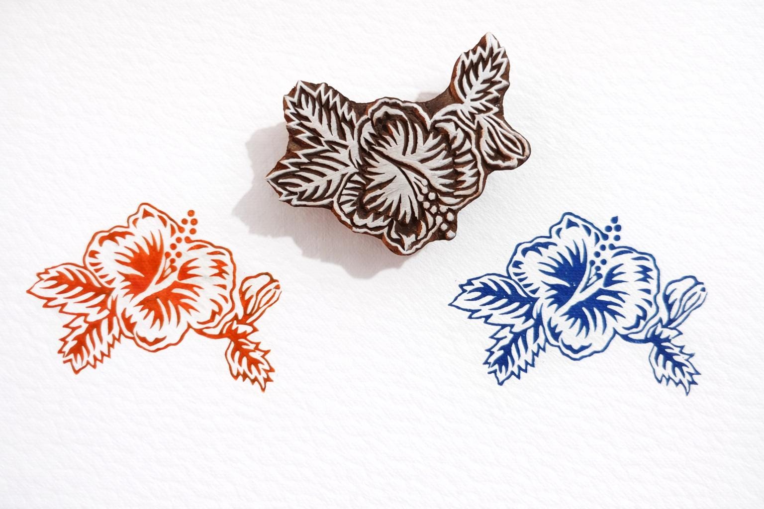 Hand carved wooden hibiscus block printing stamp creating red and blue hibiscus prints on white paper.