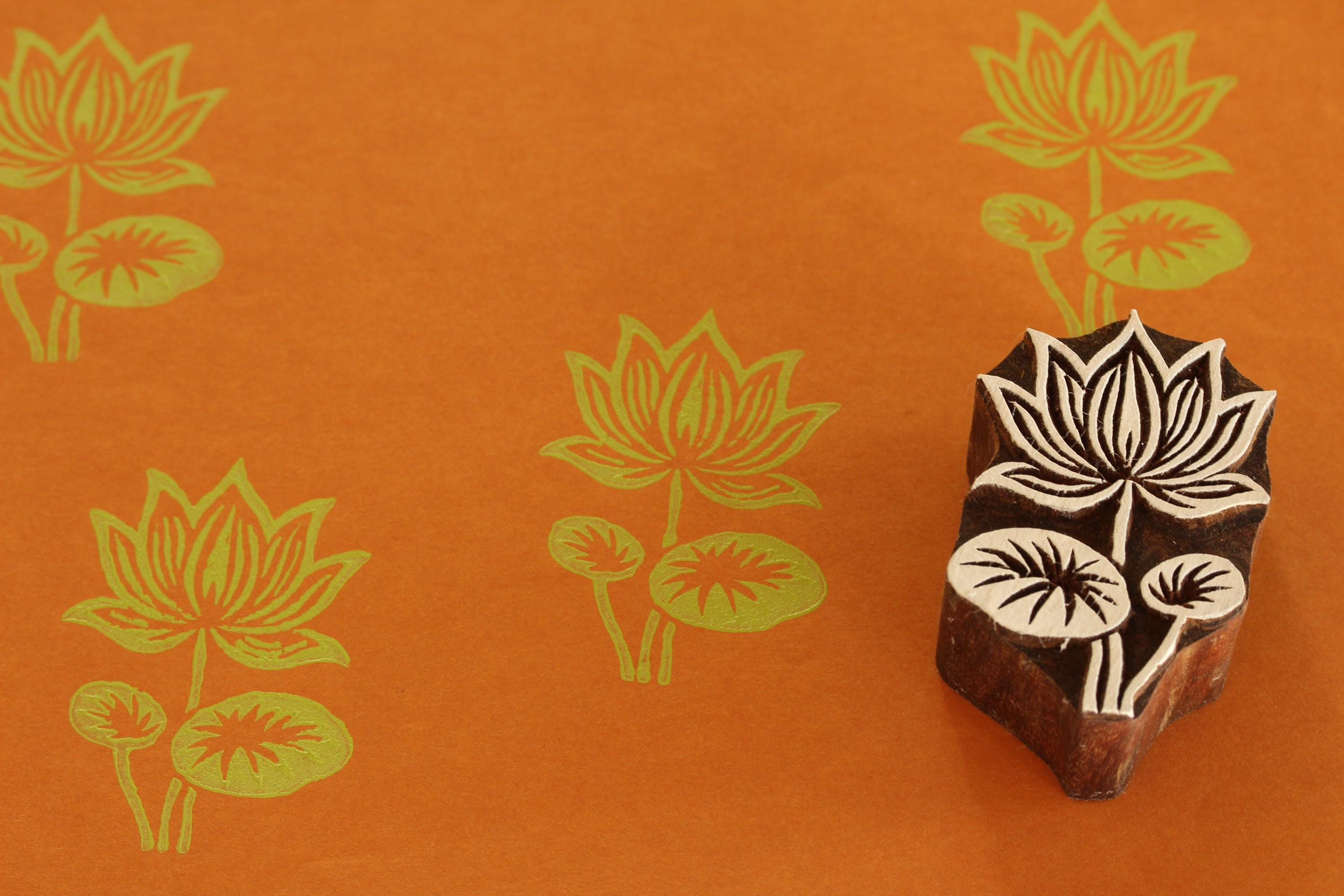 Large hand carved Indian wooden lotus printing block shown beside green lotus prints on orange paper background.